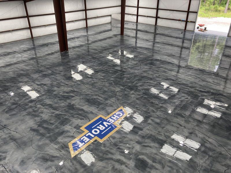 Metallic gray epoxy floor with a Chevrolet logo inlayed. The space is an empty warehouse with bright window reflections.