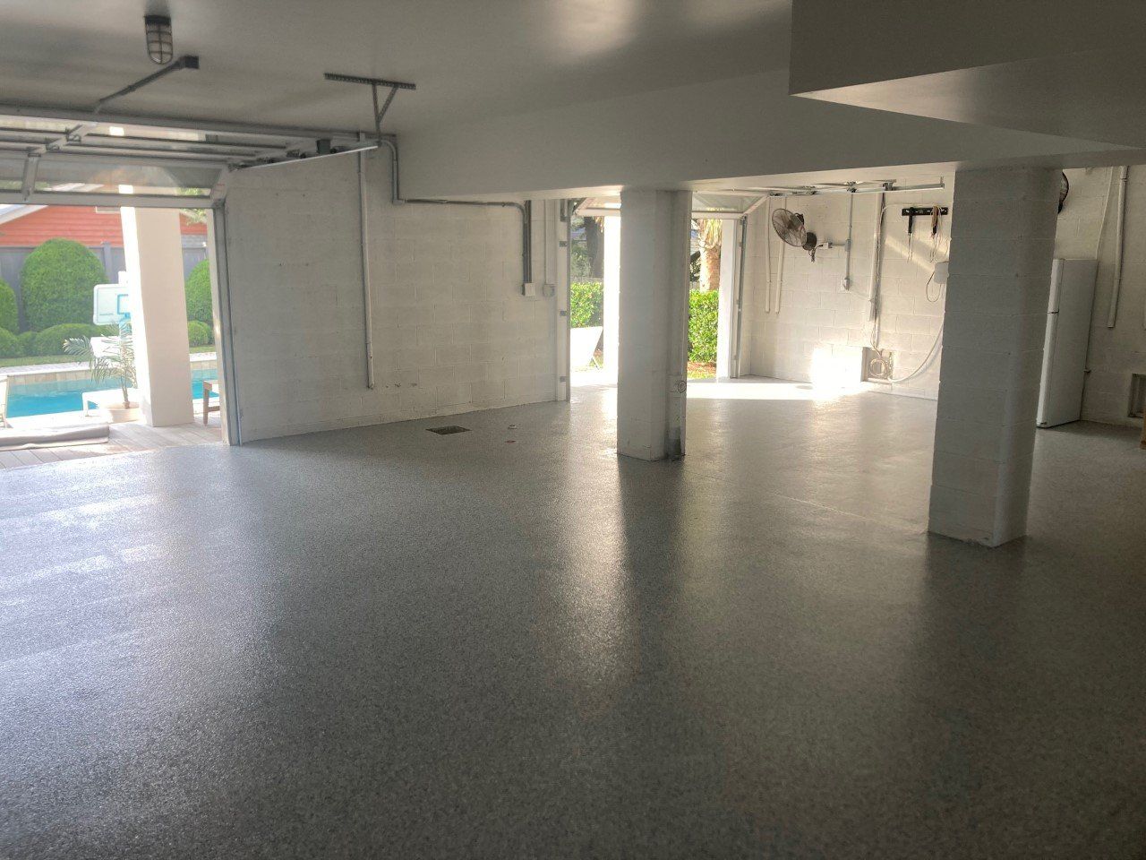 Open, concrete-floored space, possibly a garage or basement.  White walls and columns, with a view of a pool outside.