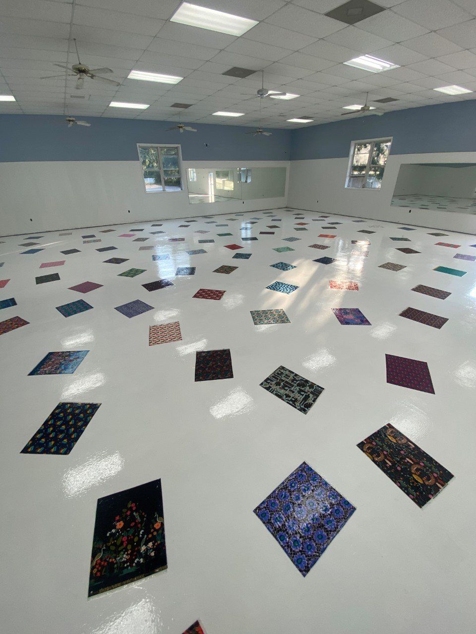 Empty room with white walls and floor, scattered colorful patterned mats. Windows are visible.