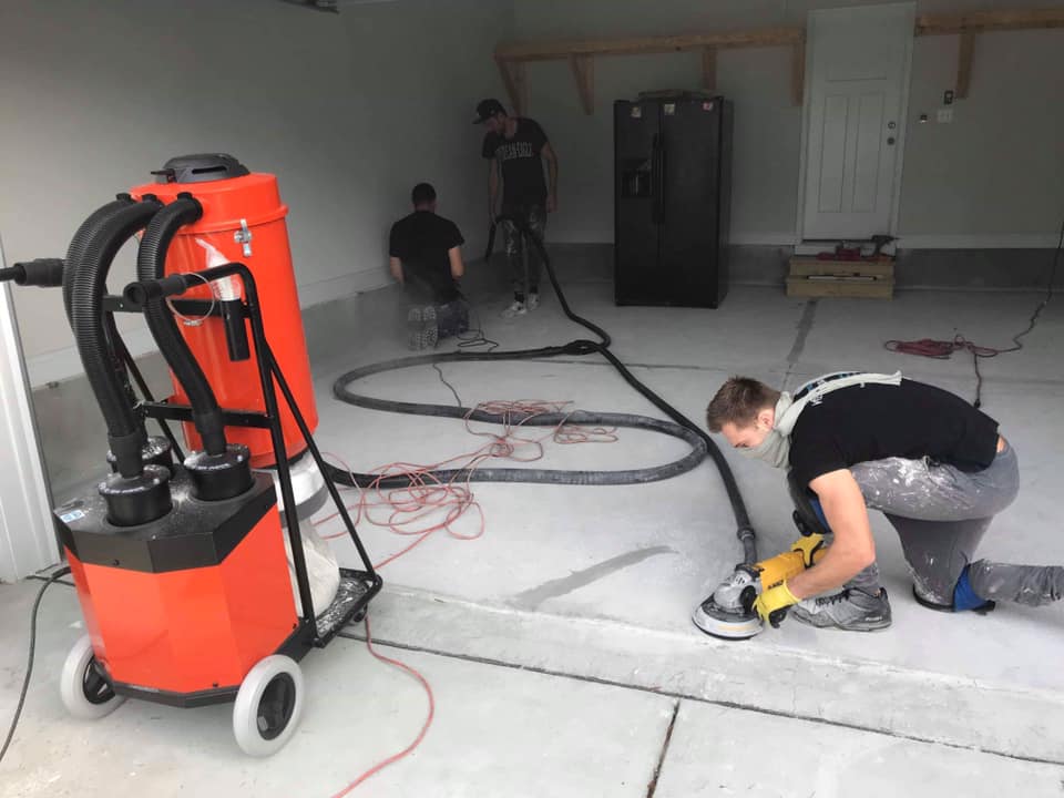 Workers grinding a concrete garage floor, connected to a large orange dust collection vacuum.