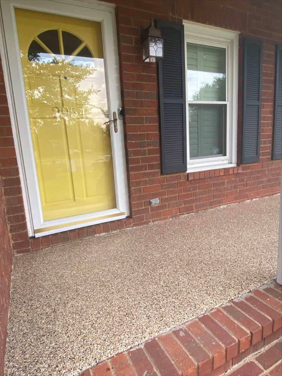 Yellow front door and brick porch with a stone-like textured surface. Next to a window with black shutters.