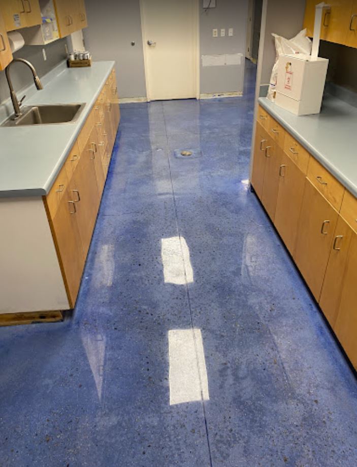 Blue polished concrete floor in a room with wood cabinets and light blue countertops. A sink and paper dispenser are visible.