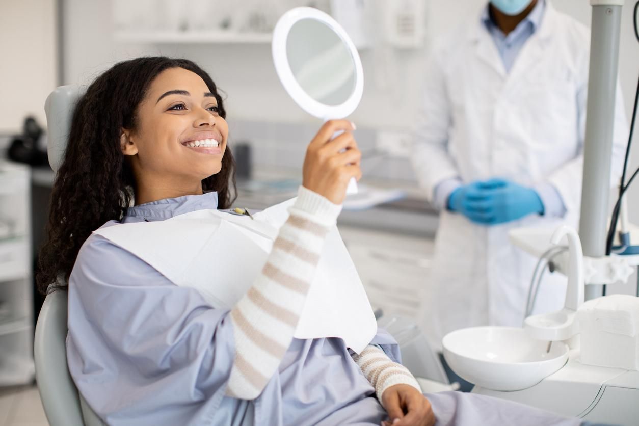 A woman is sitting in a dental chair looking at her teeth in a mirror.