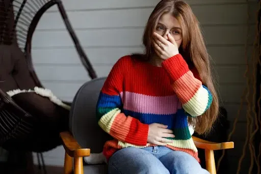 A woman in a rainbow sweater is sitting in a chair covering her mouth with her hand.