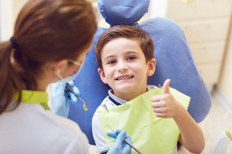 A young boy is sitting in a dental chair giving a thumbs up.