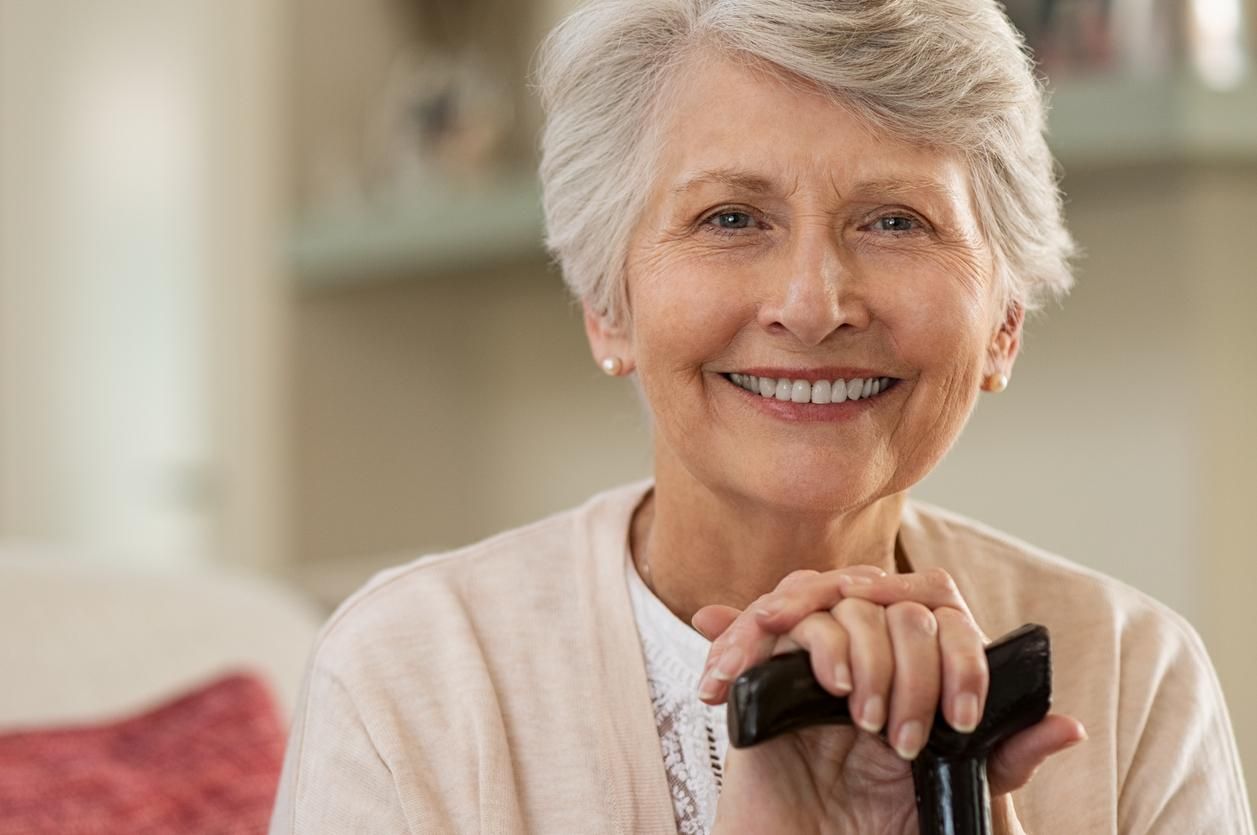 An elderly woman is sitting on a couch holding a cane and smiling.