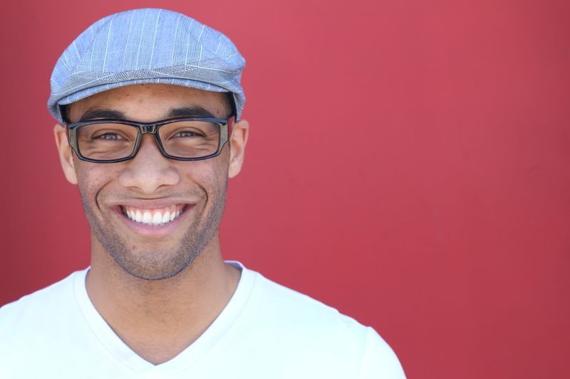 A man wearing glasses and a flat cap is smiling in front of a red background.
