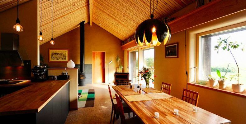 Interior of a home made with straw bale walls, dining table in the foreground, natural earth colored plaster on the walls.  Natural light fills the cozy space.