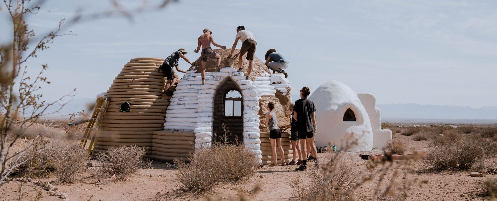 A series of earth bag domes being built in the desert by a group of people. In the background is a white completed dome with an arched window.  In the foreground workshop participants are climbing not the dome and spreading brown mud over white earth bags.