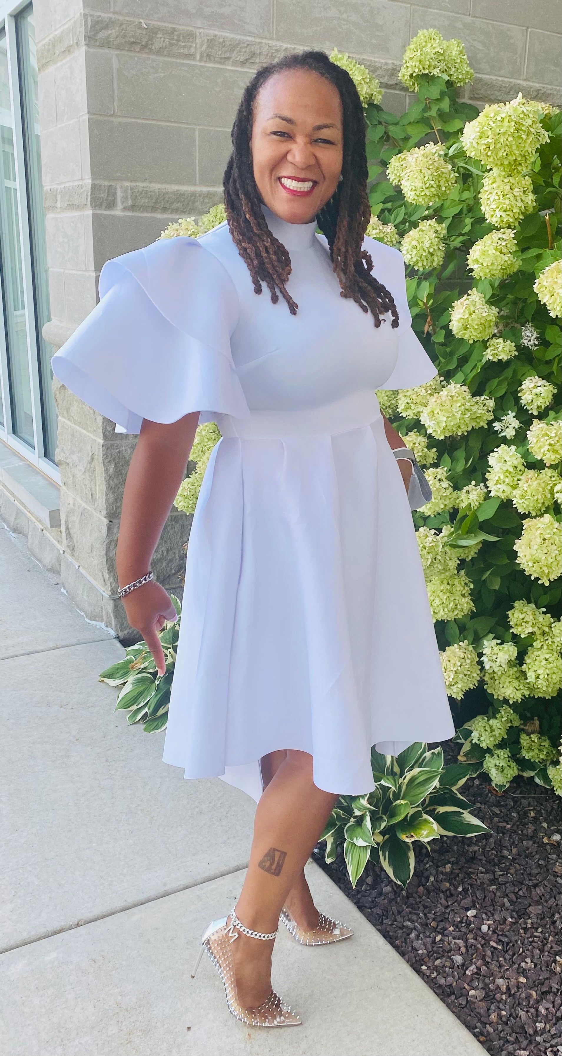 A woman in a white dress is standing on a sidewalk in front of flowers.