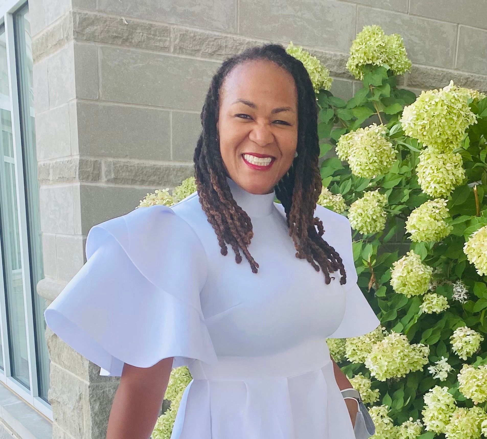 A woman in a white dress is smiling in front of a bush of flowers.