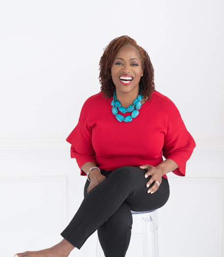 A woman in a red sweater and black pants is sitting on a clear stool and smiling.