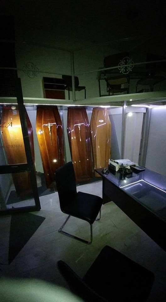 Sala espositiva di un'agenzia di pompe funebri con diverse bare in legno lucidato esposte sotto luci intense. In primo piano, una scrivania e una sedia.