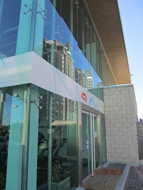 Entrada de cristal al edificio con ventanas azules reflectantes y un toldo blanco, vista desde el exterior.