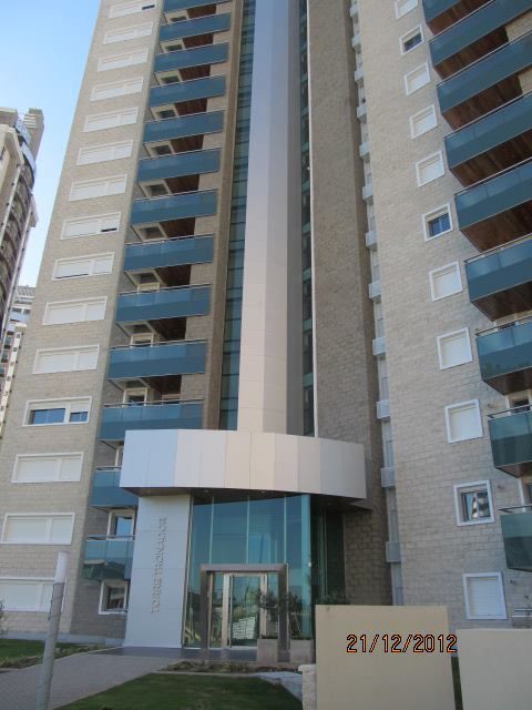 Entrada a un edificio de apartamentos alto con puertas de cristal y balcones azules bajo una marquesina blanca curva.