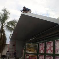 Una persona está agachada en el tejado de una tienda, con palmeras y carteles debajo.
