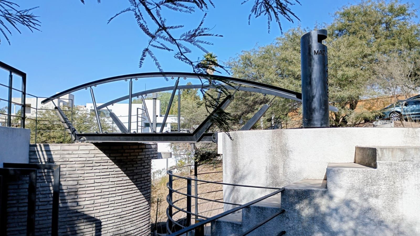 Puente peatonal moderno con barandillas metálicas sobre un canal, junto a escalones y una pared blanca bajo el cielo azul.