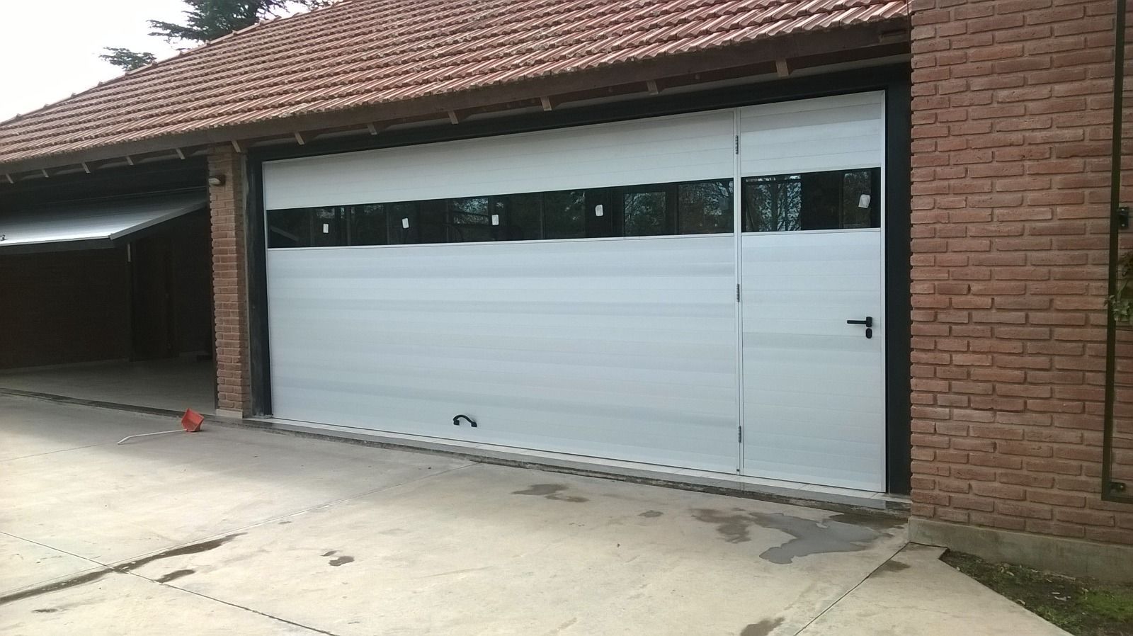 Puerta de garaje blanca en una casa de ladrillo con una estrecha franja de ventanas y entrada para vehículos al frente.