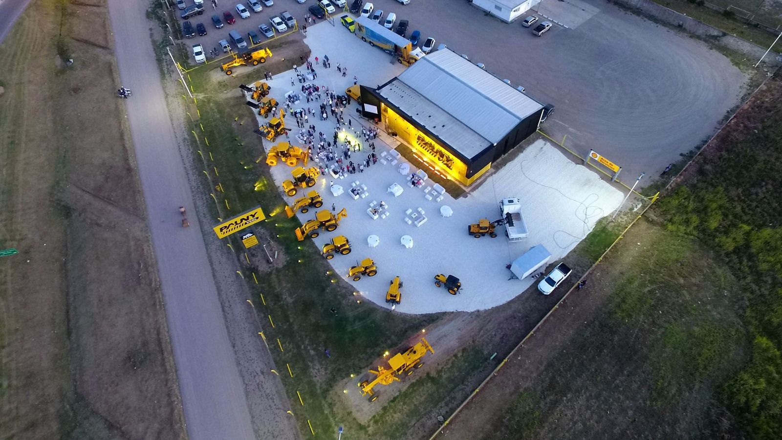 Vista aérea de una obra en construcción amarilla con equipos, vehículos y trabajadores junto a un edificio y una carretera.