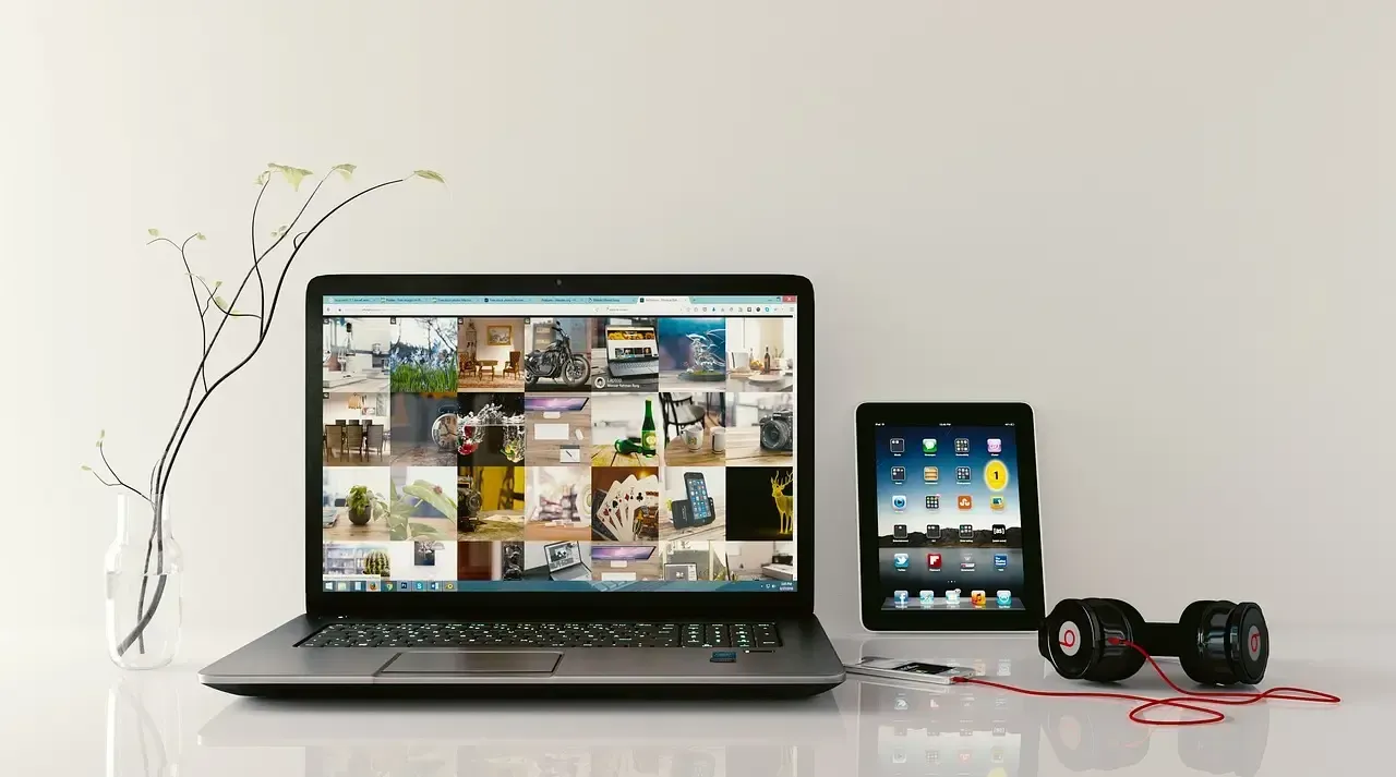 Laptop, tablet, and headphones on a white desk with a vase of flowers. The laptop screen displays multiple image previews.