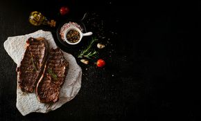 Two cooked steaks on a stone slab, with herbs, spices, and oil on a black background.