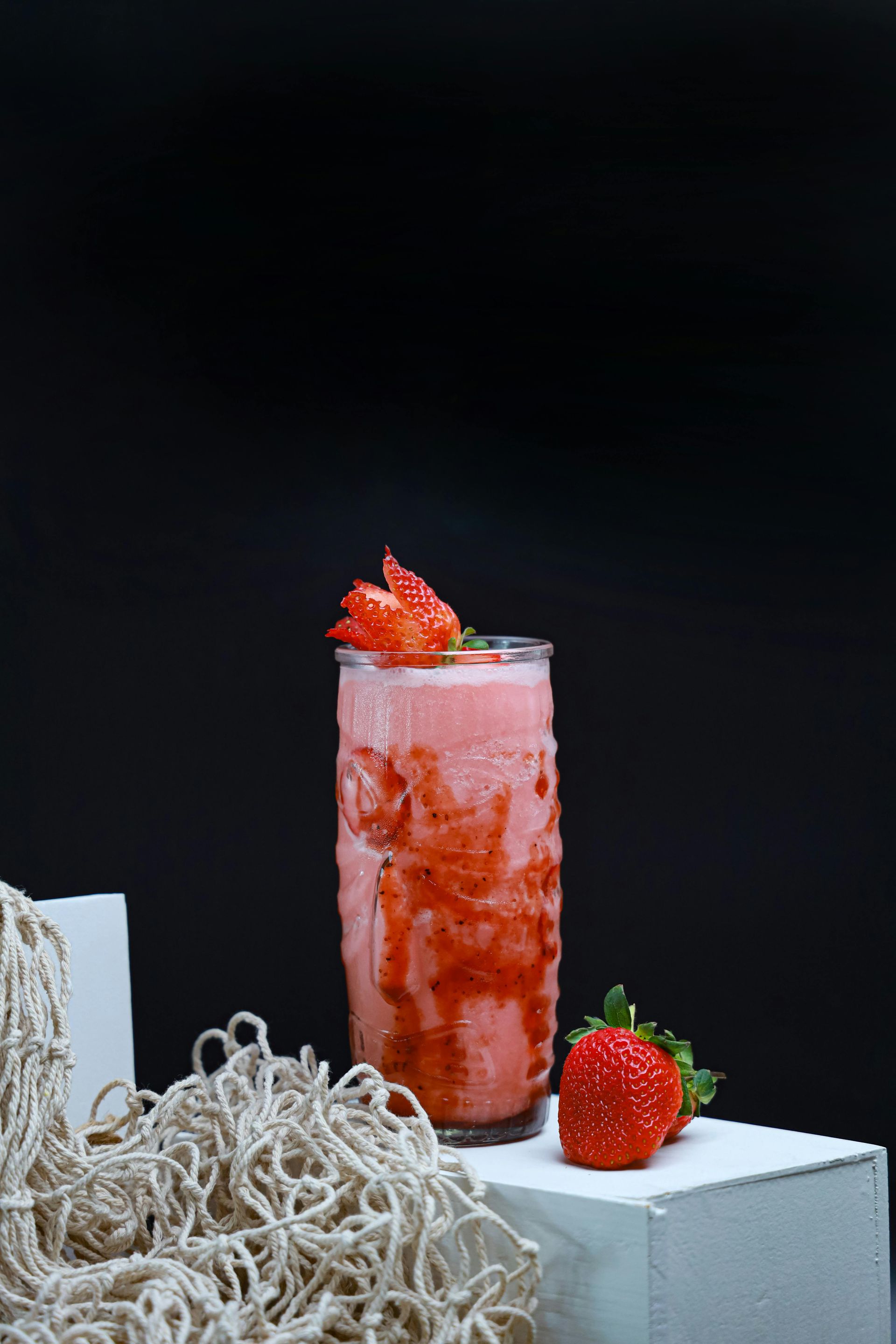 Strawberry smoothie in a glass, with a strawberry garnish and fresh strawberry on a white block, against a black backdrop.