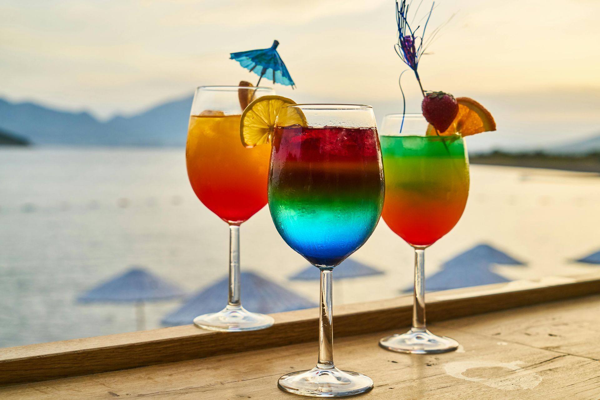 Three colorful cocktails on a wooden surface, with a beach and mountains in the background.