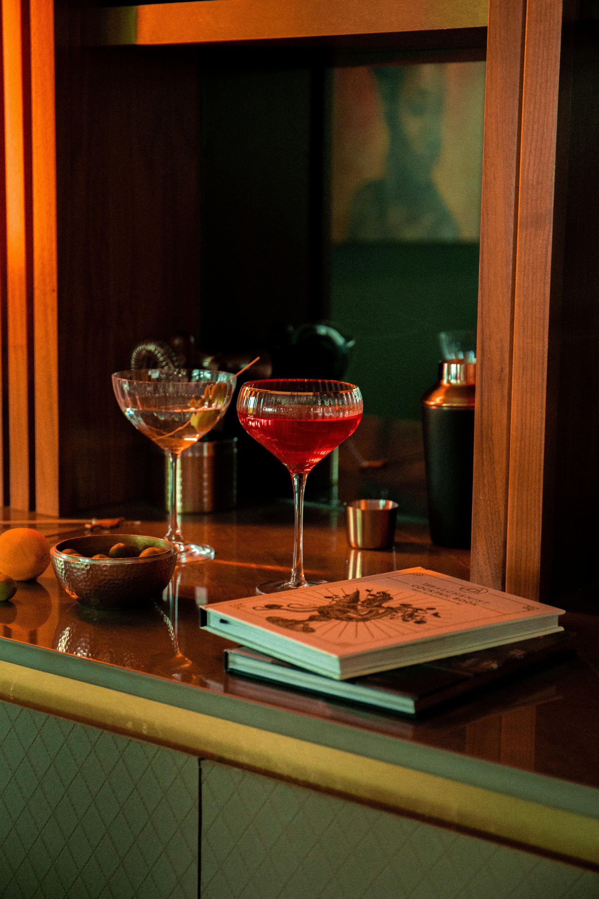 Two cocktails on a bar next to a small bowl and book. Dark setting with orange and green light.