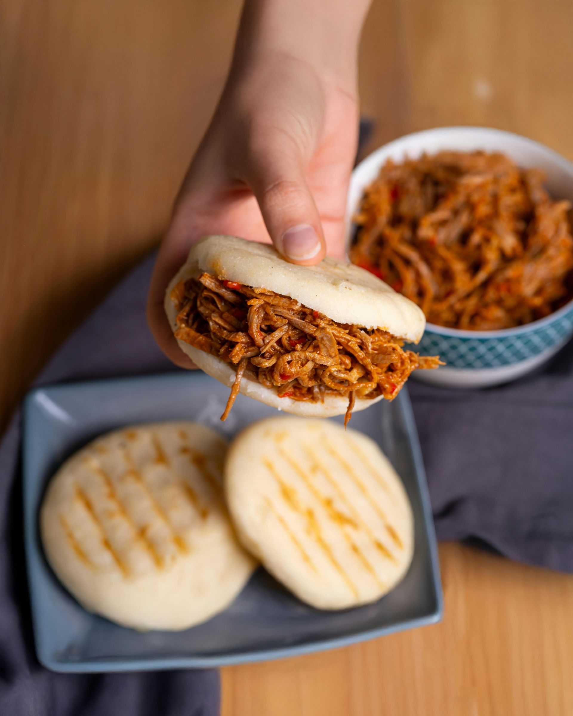 Person holding a filled, round sandwich with shredded meat. Two similar sandwiches on a plate. Bowl of meat in background.
