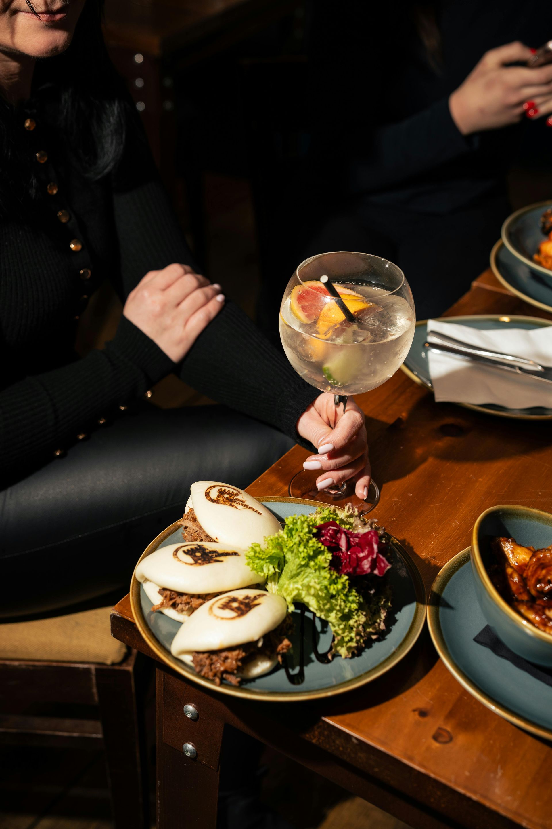 Person holding a drink next to a plate of bao buns, other dishes, on a wooden table.