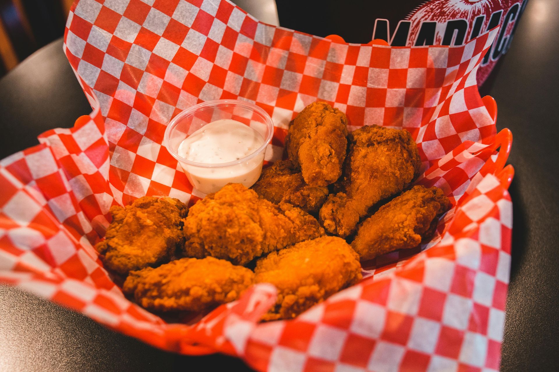 Fried chicken wings with dipping sauce in a red and white checkered basket.