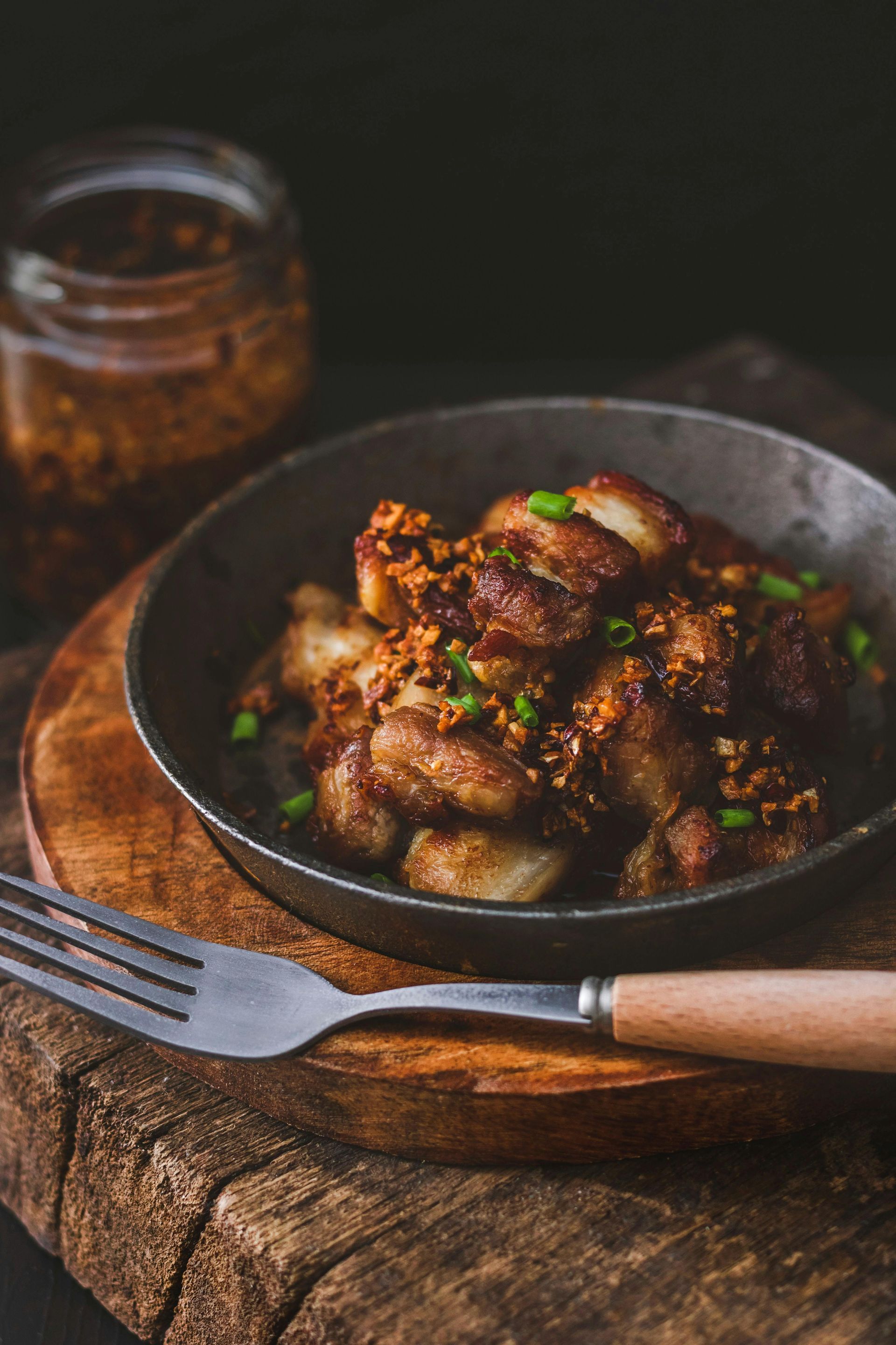 Pan of garlic-covered pork belly on a wooden board, with a jar of sauce and a fork nearby.