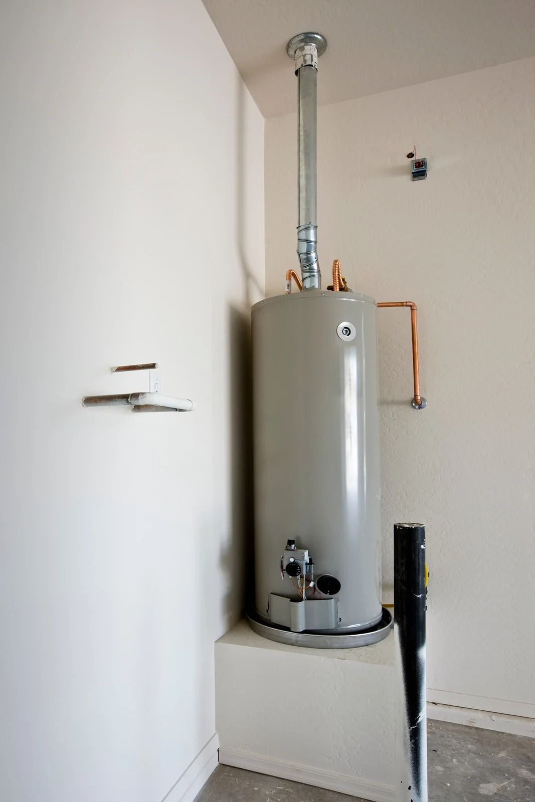Gray water heater in a corner basement room with exposed pipes and a black umbrella leaning nearby