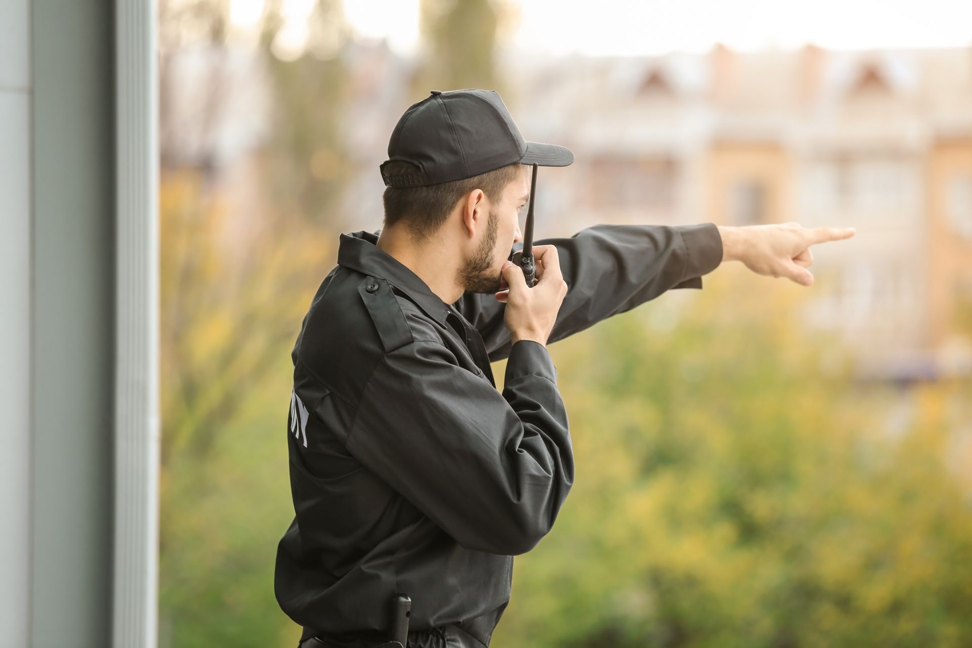 Person in black cap and jacket points outdoors while speaking into a radio.