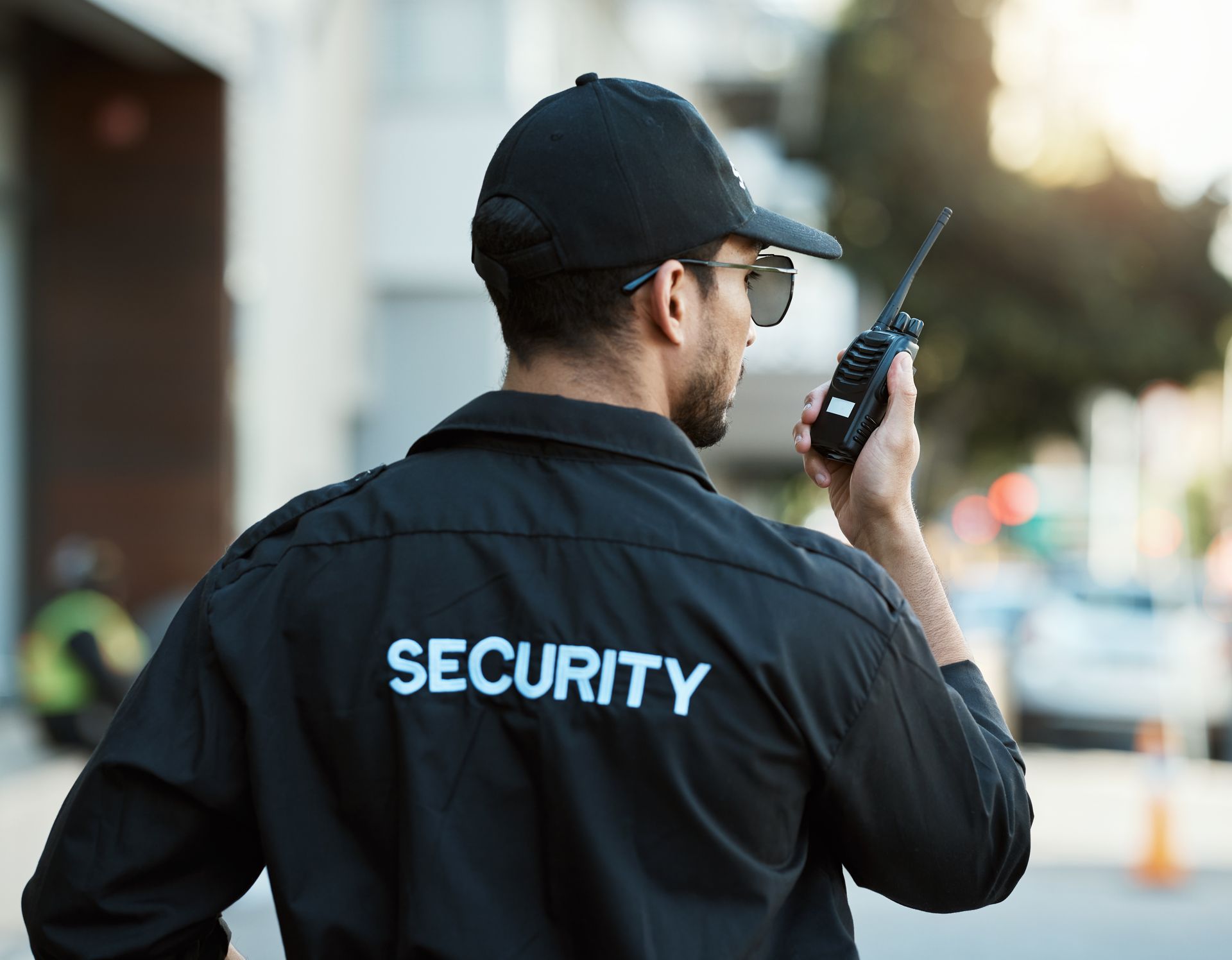 Security guard in black jacket and cap, holding a walkie-talkie on a city street.