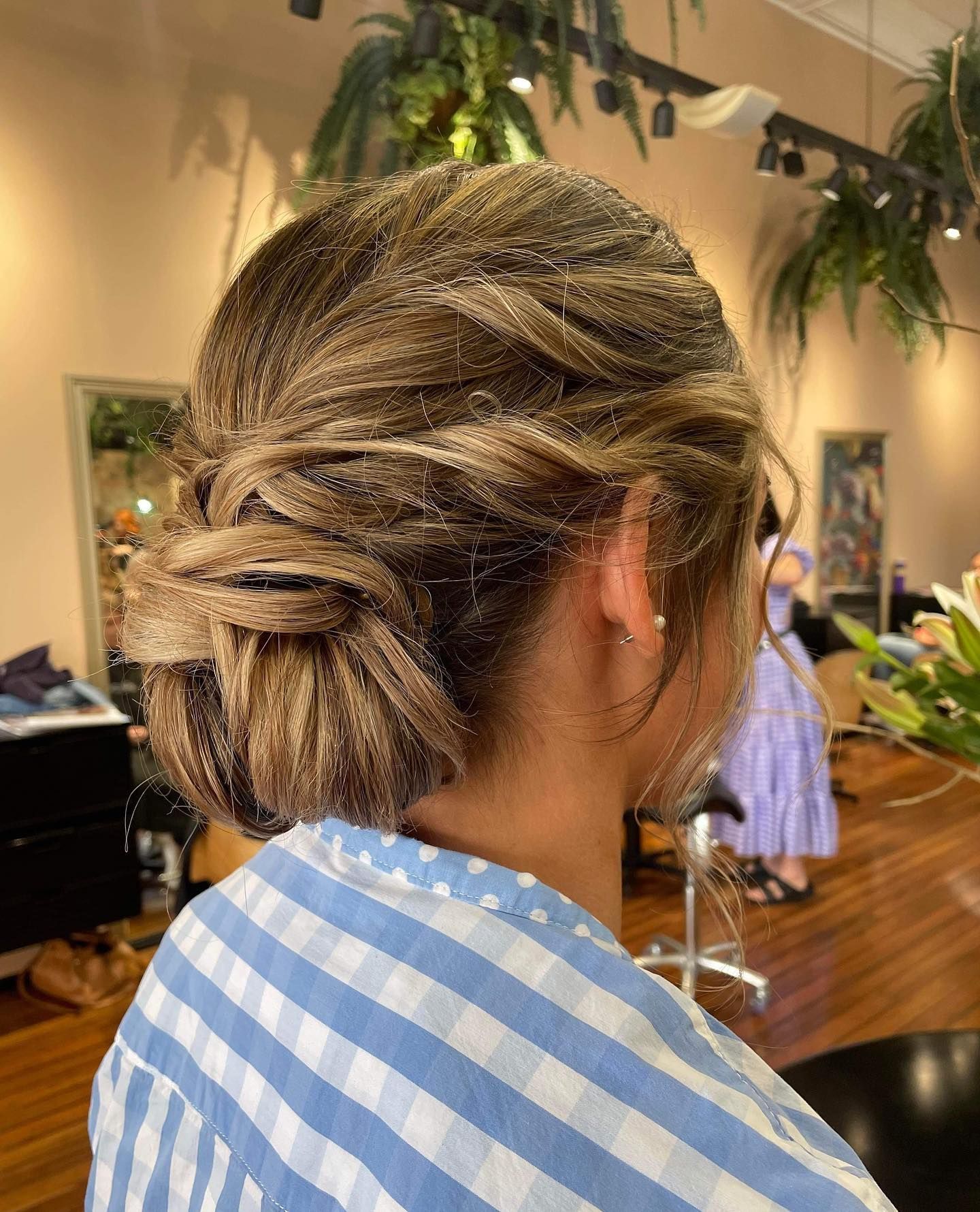 Woman with blonde-brown hair styled in a low twisted bun, wearing a blue and white checked shirt.