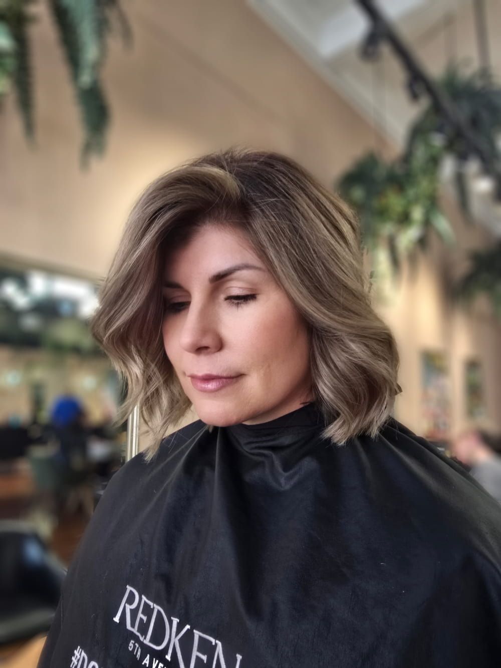 Woman with highlighted bob haircut in salon, wearing a Redken cape.