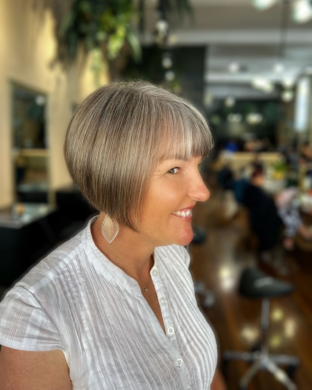 Woman with a short, gray bob hairstyle smiles in a salon. She wears a white shirt and earrings.