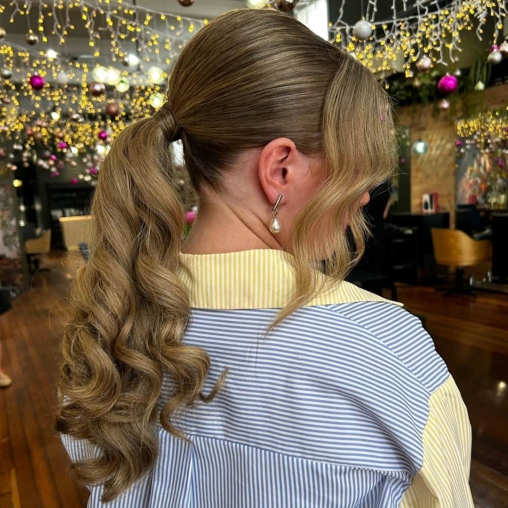 Woman with wavy ponytail and side-swept bangs, wearing a blue and yellow striped shirt, in a salon.