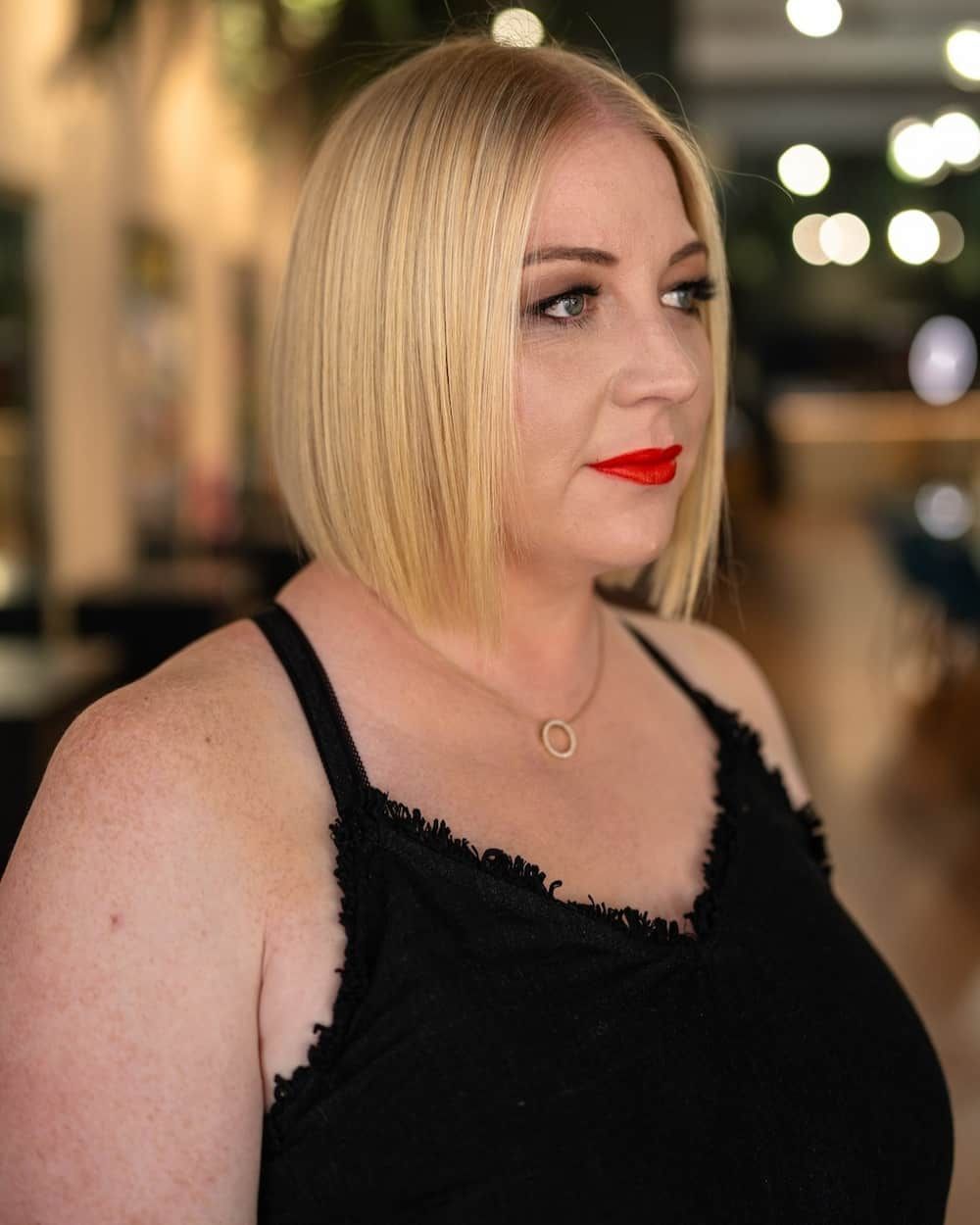 Woman with blonde bob and red lipstick, wearing a black tank top, in a well-lit room.