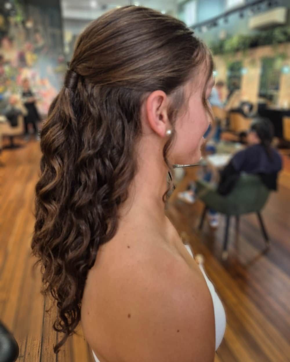 Woman with half-up curly brown hair, wearing a white top, in a salon with wooden floors.