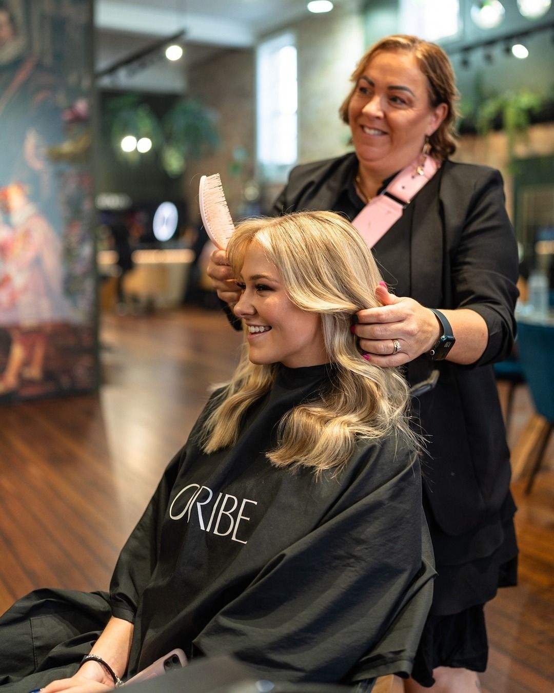 Blonde woman getting her hair styled by a stylist in a salon. Both women are smiling.
