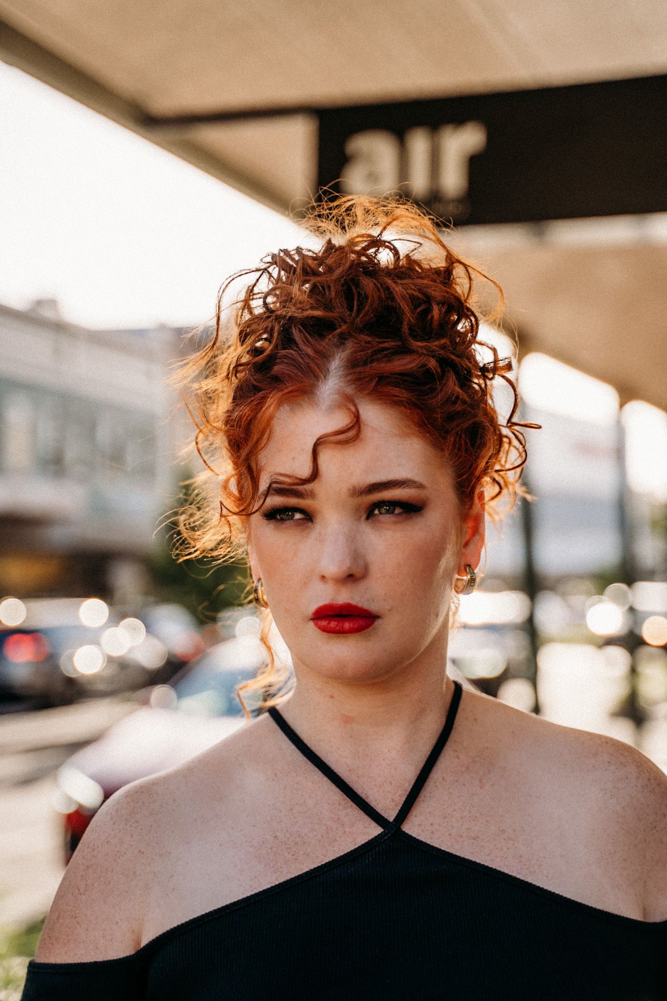 Woman with red hair and lipstick, wearing a black top. Standing outside near a sign that says
