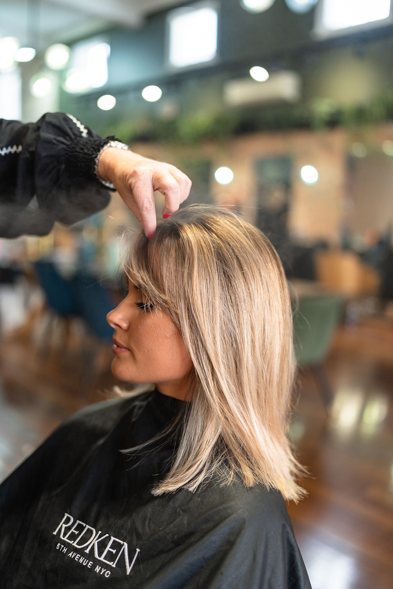 Woman getting her hair styled at a salon; highlights, blunt bangs, black salon cape, stylist's hand.