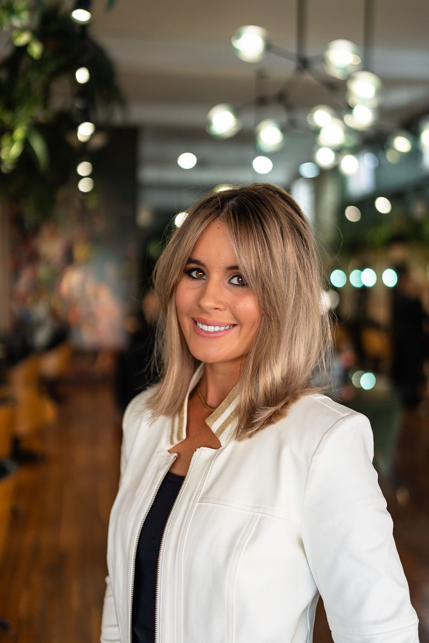 Woman with blonde bob, smiling, in white jacket, indoors with lights and greenery.