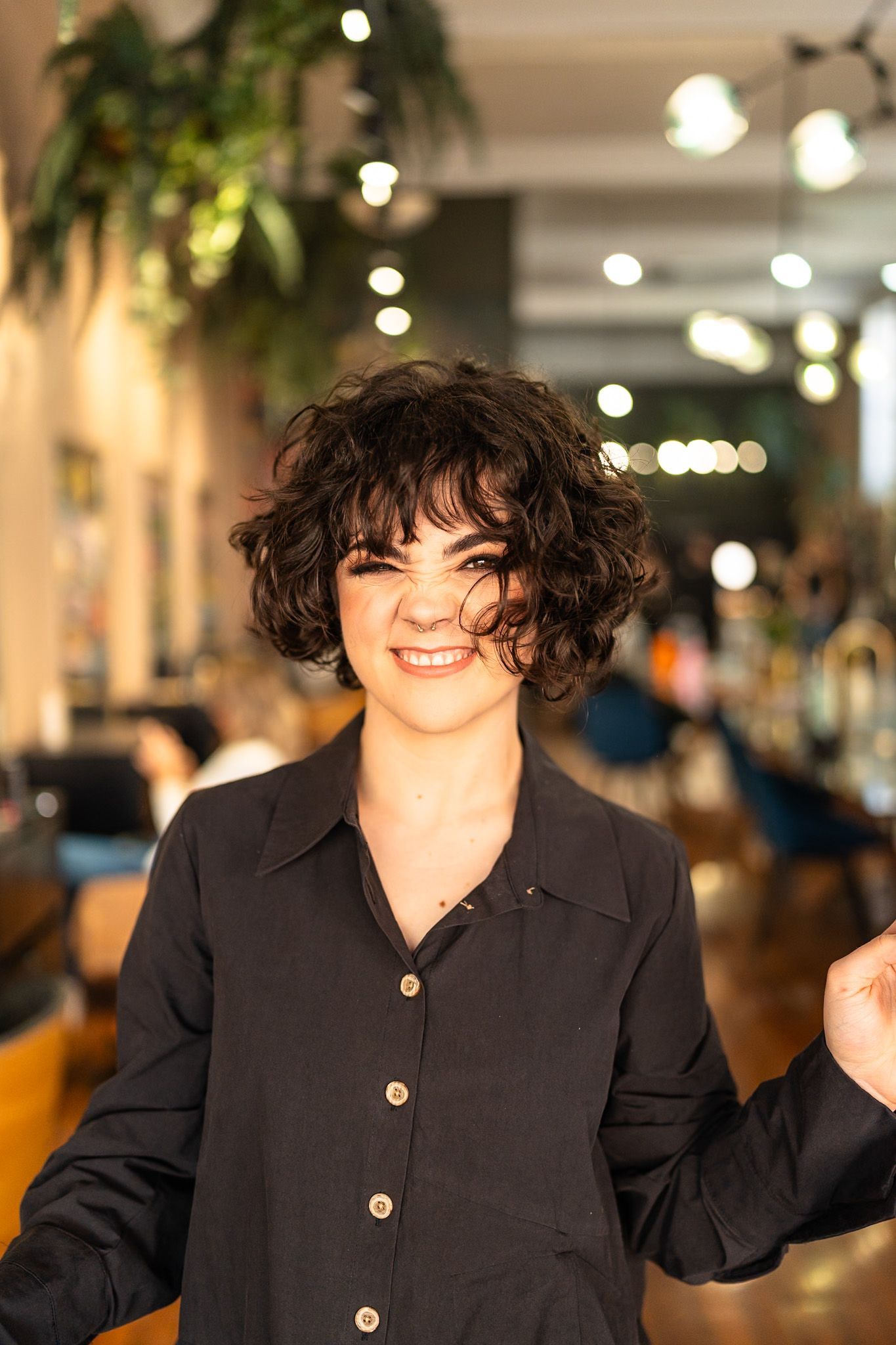 Woman with curly dark hair smiles, wearing a black button-down shirt, in a salon.