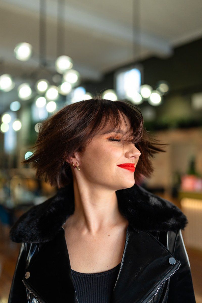 Woman with red lipstick and short brown hair, wearing a black jacket, indoors with soft lights.