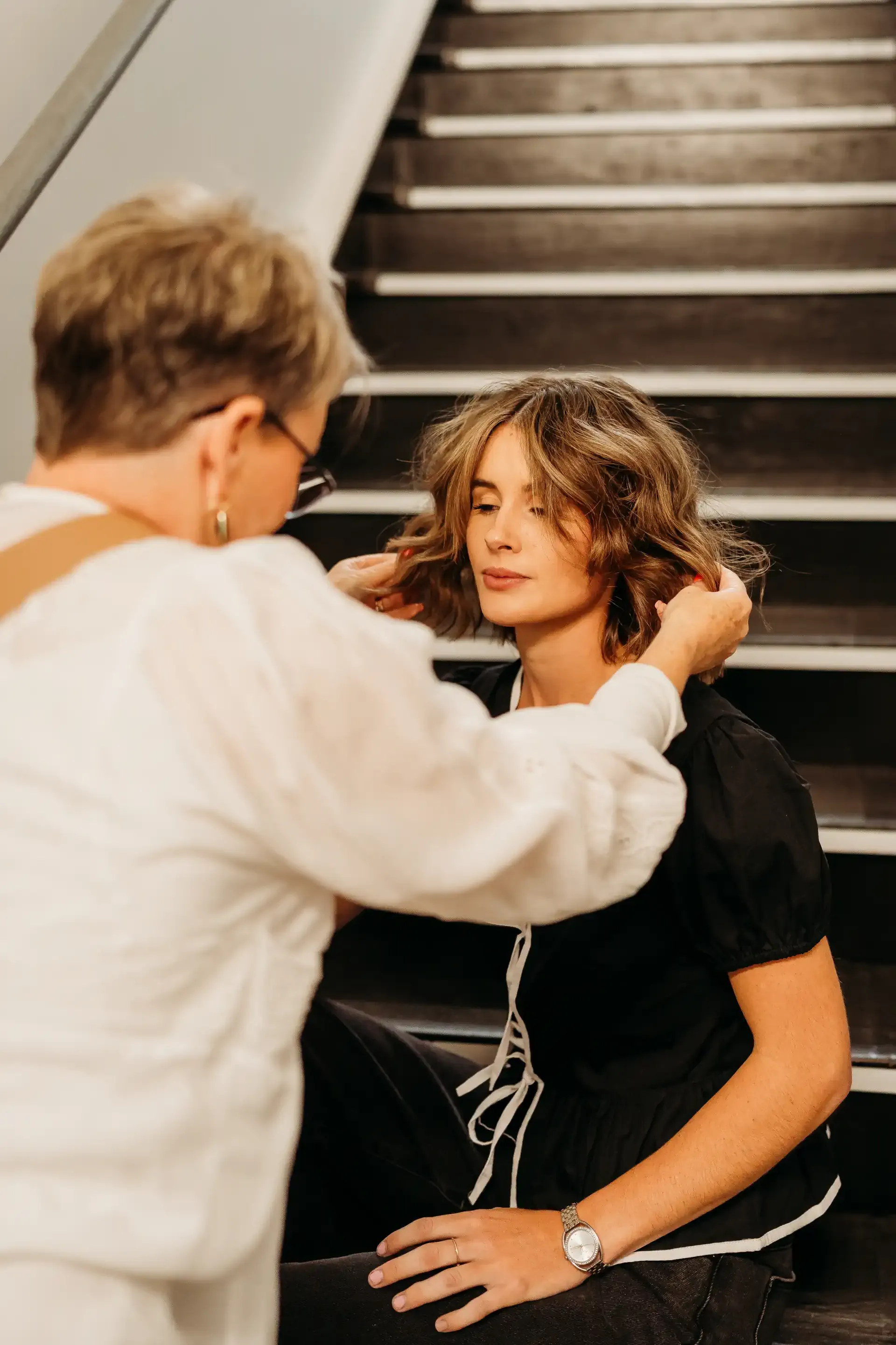A woman is sitting on a staircase getting her hair done by a hairdresser.