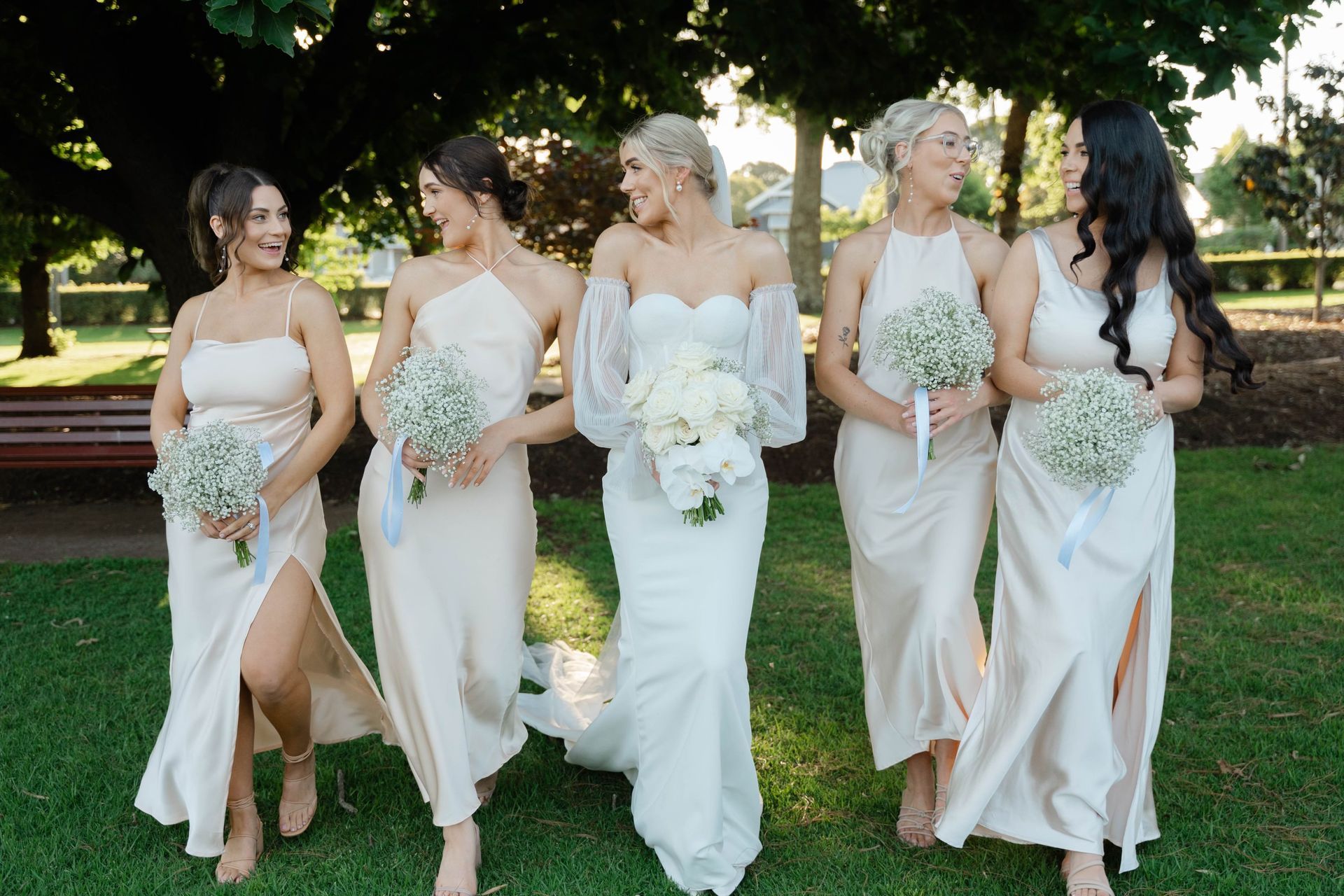 Bride and bridesmaids in a park, smiling. Bride wears a white gown; bridesmaids in light peach dresses.