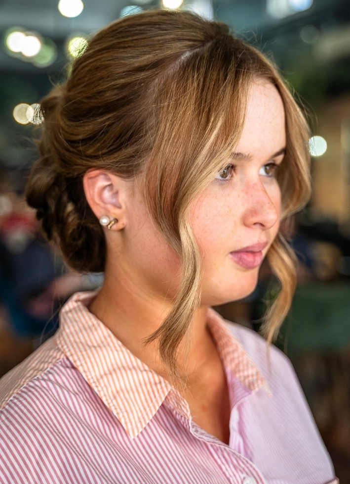 Woman with light brown hair styled in an updo and face framing waves, wearing a pink pinstripe shirt.